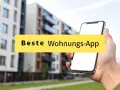 Hand hält Smartphone mit der Aufschrift "Beste Wohnungs-App" vor modernen Gebäuden.