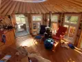 Gemütliches Inneres einer Jurte als Wohnhaus mit Holzofen, Küche und Sitzgelegenheiten. Sonnenlicht fällt durch die Fenster.