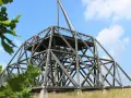 Der Spurwerkturm Waltrop, eine beeindruckende Holzkonstruktion, ragt gegen einen blauen Himmel mit Wolken empor. Grüne Blätter rahmen die Szene ein.