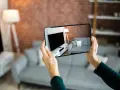Hände halten Smartphone, das eine Wohnung mit Sofa und Fernseher zeigt. Darf ein Makler Fotos von meiner Wohnung machen? Ja, für die Vermarktung.