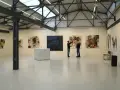 Zwei Personen betrachten Kunstwerke in der Kunsthalle Burkamp. Abstrakte Gemälde hängen an den weißen Wänden, ein Tisch mit Gläsern steht in der Mitte.