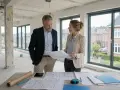 Architekten besprechen Pläne für die Umnutzung Gewerbe zu Wohnraum in einem Rohbau mit großen Fenstern und Blick auf Häuser.