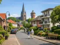 Idyllische Straße in Porta Westfalica mit Kirche und Wasserturm. Ein älteres Paar spaziert, während Autos parken. Der Mietspiegel Porta Westfalica spiegelt diese ruhige Wohngegend wider.