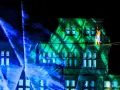 Ein Artist balanciert auf einem Hochseil vor einem Gebäude, das mit grünen Lichtmustern beleuchtet ist. Blaue Lichtstrahlen durchziehen die Szene. Das ist ein Highlight, wann ist Recklinghausen leuchtet!