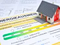 Modellhaus auf einem Energieausweis (Verbrauchsausweis oder Bedarfsausweis). Der Ausweis zeigt den Energiebedarf eines Gebäudes mit einer Skala von grün bis rot.