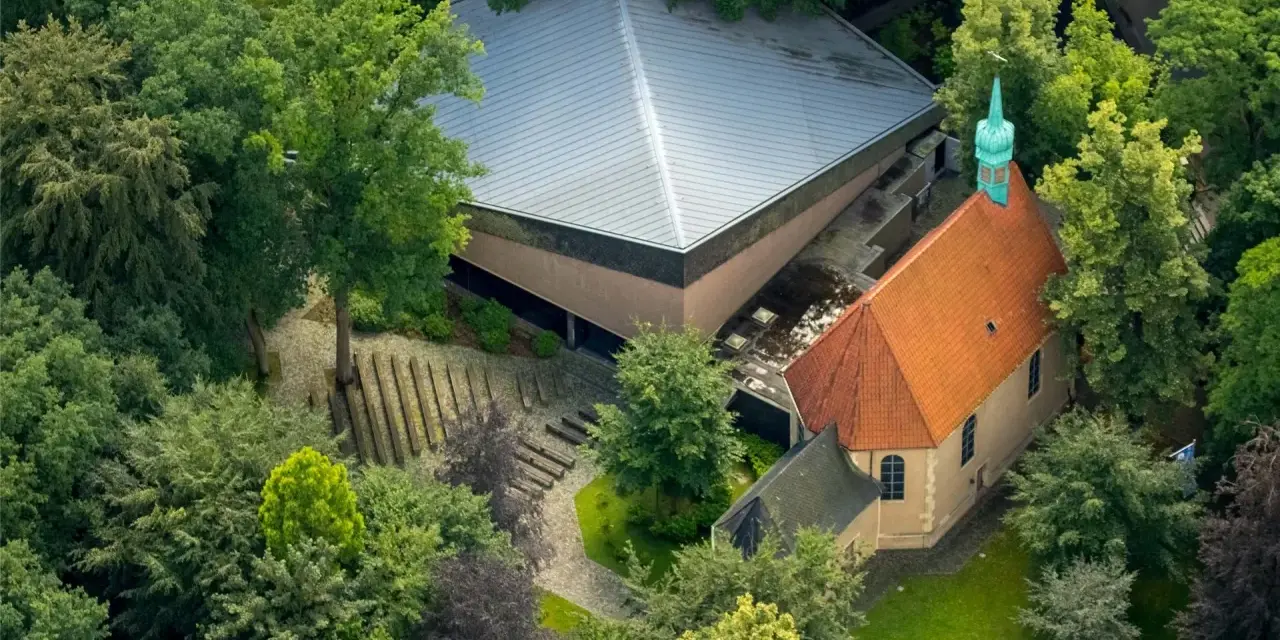 Luftaufnahme von Annaberg Haltern: Ein Gotteshaus mit rotem Ziegeldach und grüner Turmspitze, daneben ein moderner Bau mit Pyramiden-Dach, umgeben von üppigem Grün.