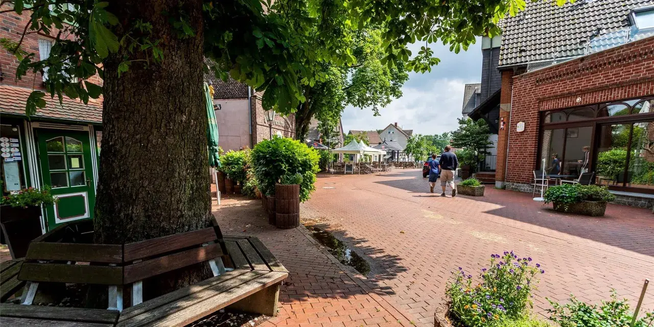 Ein lauschiger Kirchplatz in Recklinghausen mit Kopfsteinpflaster, Bäumen und Sitzgelegenheiten. Ein Paar spaziert vorbei an Cafés mit Außenbestuhlung.