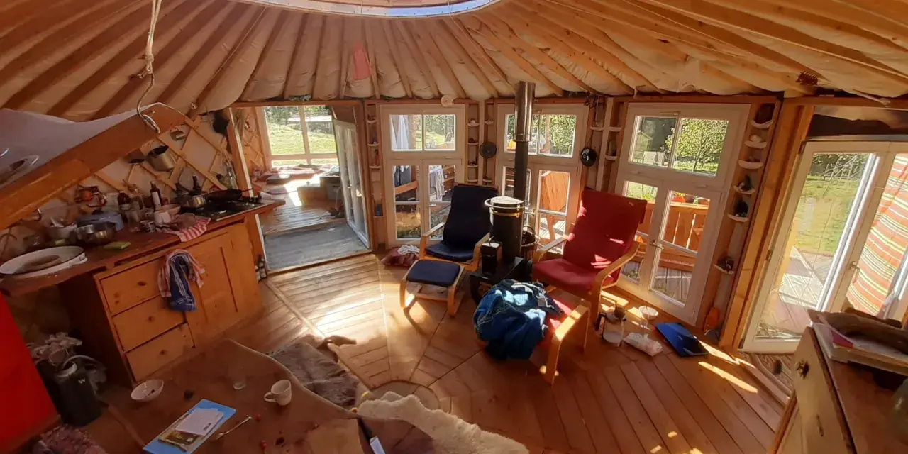 Gemütliches Inneres einer Jurte als Wohnhaus mit Holzofen, Küche und Sitzgelegenheiten. Sonnenlicht fällt durch die Fenster.