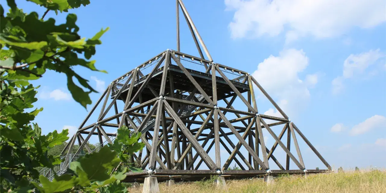 Der Spurwerkturm Waltrop, eine beeindruckende Holzkonstruktion, ragt gegen einen blauen Himmel mit Wolken empor. Grüne Blätter rahmen die Szene ein.