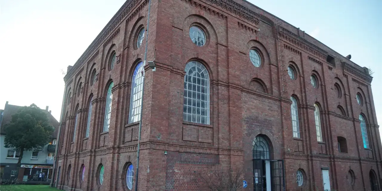 Das imposante Backsteingebäude der Zeche Scherlbeck mit seinen vielen Rund- und Bogenfenstern, einige davon bunt beleuchtet, strahlt historischen Charme aus.