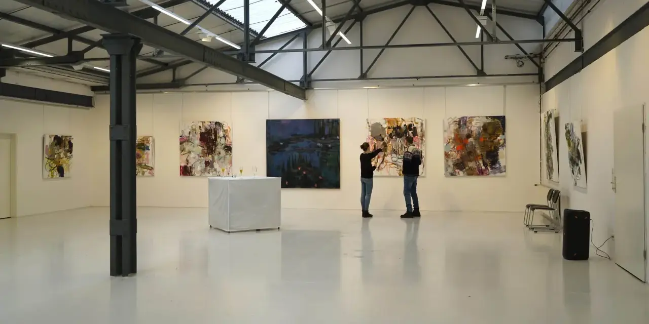 Zwei Personen betrachten Kunstwerke in der Kunsthalle Burkamp. Abstrakte Gemälde hängen an den weißen Wänden, ein Tisch mit Gläsern steht in der Mitte.