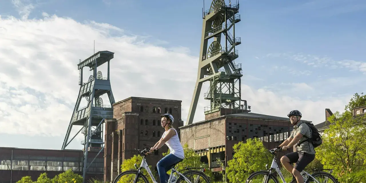 Ein Paar genießt eine Fahrradtour in Recklinghausen, vorbei an alten Fördertürmen und Industriegebäuden.