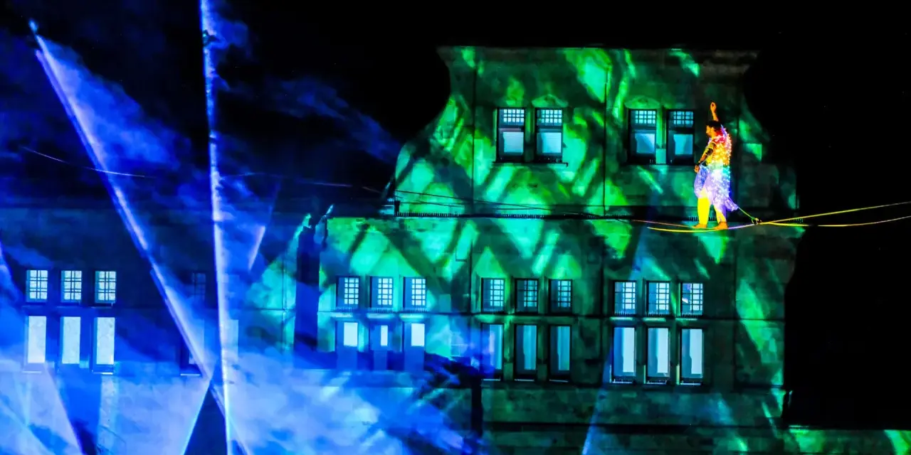 Ein Artist balanciert auf einem Hochseil vor einem Gebäude, das mit grünen Lichtmustern beleuchtet ist. Blaue Lichtstrahlen durchziehen die Szene. Das ist ein Highlight, wann ist Recklinghausen leuchtet!