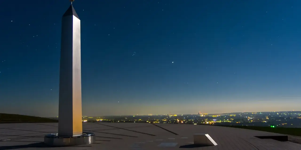 Die Sonnenuhr in Herten ragt unter einem sternenklaren Nachthimmel empor. Ein glänzender Obelisk wirft lange Schatten auf die gepflasterte Fläche.