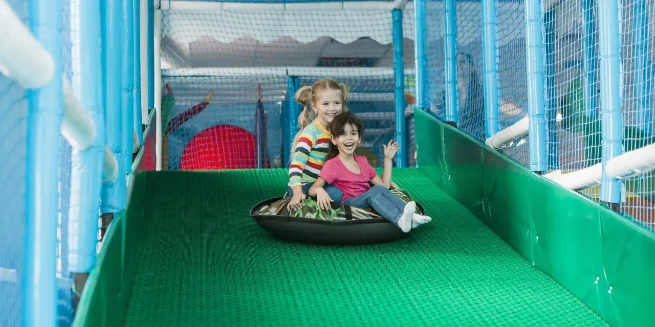 Zwei lachende Mädchen rutschen auf einem Schlauchboot in einem Indoor-Spielplatz.