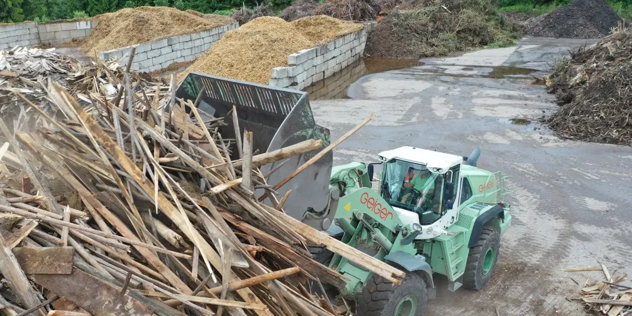 Radlader sammelt Holzreste für die Wiederverwertung. Große Haufen von recycelten Materialien sind im Hintergrund zu sehen.