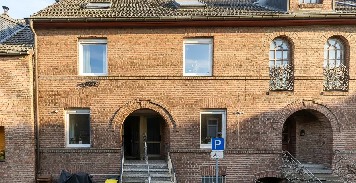 Ziegelhaus mit mehreren Fenstern und Eingängen. Ein Parkplatzschild weist auf die Lage hin, die für die Neuss Immobilienpreise relevant ist.