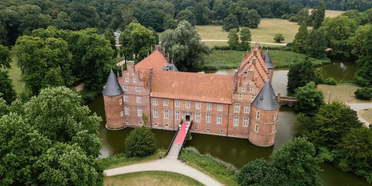 Schloss Recklinghausen, ein imposantes Wasserschloss, umgeben von grünen Bäumen und einem Teich. Eine rote Teppich führt zur Eingangstür.