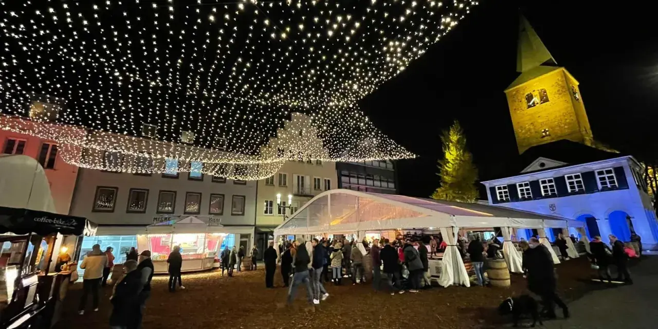 Glitzernde Lichterketten schmücken den **Weihnachtsmarkt in Dorsten**. Menschen bummeln unter Zelten und vor Buden, während im Hintergrund die beleuchtete Kirche thront.