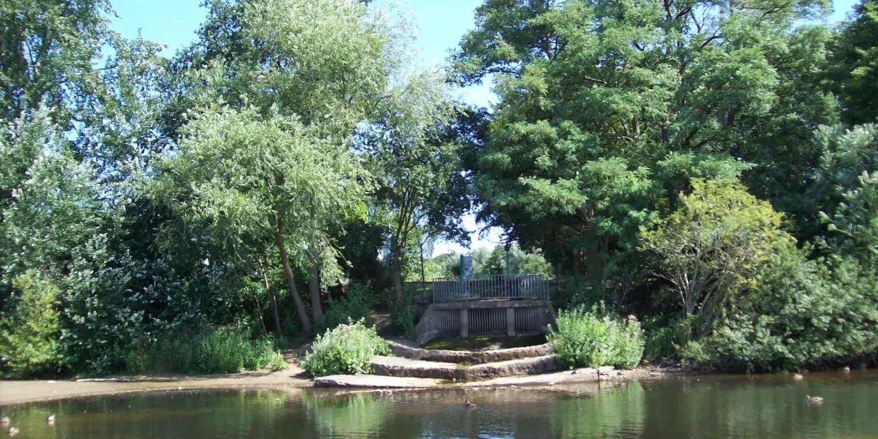 Ein ruhiger blauer See in Dorsten, umgeben von üppigem Grün. Eine kleine Holzbrücke überspannt das Wasser, während Enten friedlich schwimmen.