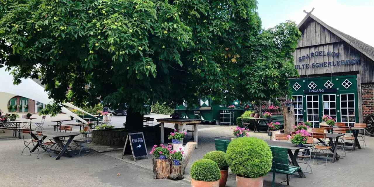 Einladende Sitzgelegenheiten unter einem großen Baum im naturerlebnisgarten Herten. Der Spargelhof mit grünen Türen und der Aufschrift "Eingang" ist im Hintergrund zu sehen.
