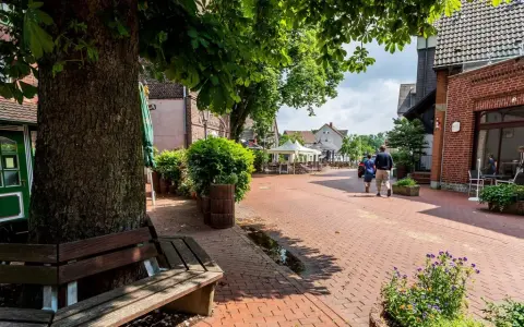 Ein lauschiger Kirchplatz in Recklinghausen mit Kopfsteinpflaster, Bäumen und Sitzgelegenheiten. Ein Paar spaziert vorbei an Cafés mit Außenbestuhlung.