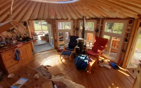 Gemütliches Inneres einer Jurte als Wohnhaus mit Holzofen, Küche und Sitzgelegenheiten. Sonnenlicht fällt durch die Fenster.