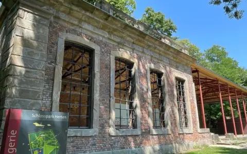 Die Orangerie Herten, ein historisches Gebäude mit großen Fensterfronten, steht im Schatten von Bäumen.