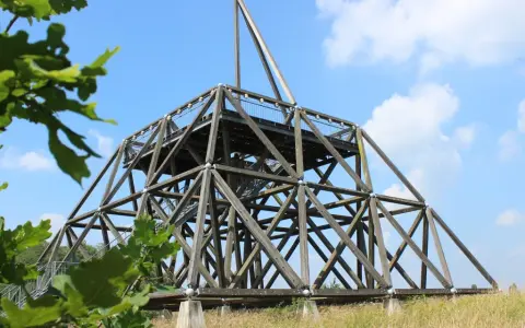 Der Spurwerkturm Waltrop, eine beeindruckende Holzkonstruktion, ragt gegen einen blauen Himmel mit Wolken empor. Grüne Blätter rahmen die Szene ein.