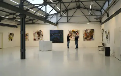 Zwei Personen betrachten Kunstwerke in der Kunsthalle Burkamp. Abstrakte Gemälde hängen an den weißen Wänden, ein Tisch mit Gläsern steht in der Mitte.