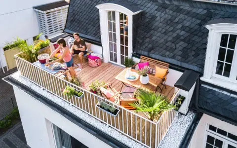 Ein Paar genießt den Sommer auf einem Balkon mit Holz-Bodenbelag, ideal für eine Mietwohnung. Grillen und Entspannen stehen im Vordergrund.