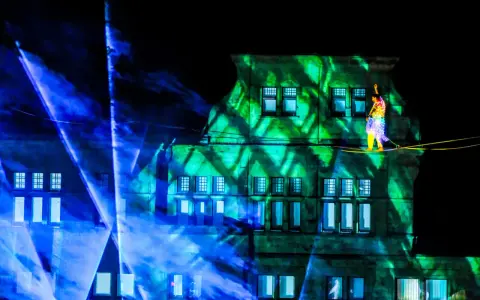 Ein Artist balanciert auf einem Hochseil vor einem Gebäude, das mit grünen Lichtmustern beleuchtet ist. Blaue Lichtstrahlen durchziehen die Szene. Das ist ein Highlight, wann ist Recklinghausen leuchtet!