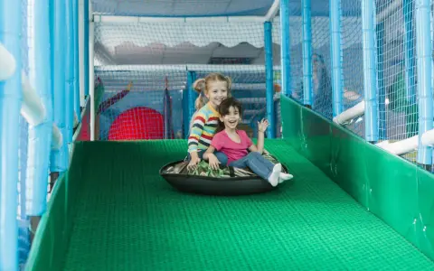 Zwei lachende Mädchen rutschen auf einem Schlauchboot in einem Indoor-Spielplatz.