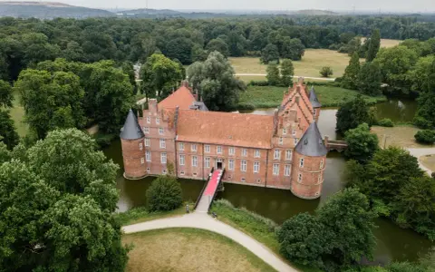 Schloss Recklinghausen, ein imposantes Wasserschloss, umgeben von grünen Bäumen und einem Teich. Eine rote Teppich führt zur Eingangstür.