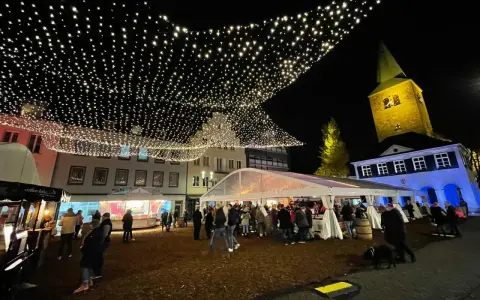 Glitzernde Lichterketten schmücken den **Weihnachtsmarkt in Dorsten**. Menschen bummeln unter Zelten und vor Buden, während im Hintergrund die beleuchtete Kirche thront.
