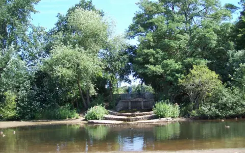 Ein ruhiger blauer See in Dorsten, umgeben von üppigem Grün. Eine kleine Holzbrücke überspannt das Wasser, während Enten friedlich schwimmen.