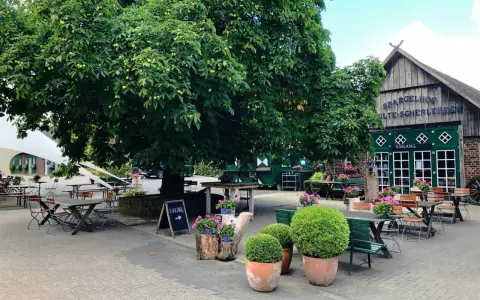 Einladende Sitzgelegenheiten unter einem großen Baum im naturerlebnisgarten Herten. Der Spargelhof mit grünen Türen und der Aufschrift "Eingang" ist im Hintergrund zu sehen.