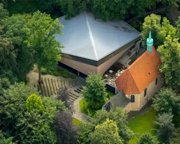 Luftaufnahme von Annaberg Haltern: Ein Gotteshaus mit rotem Ziegeldach und grüner Turmspitze, daneben ein moderner Bau mit Pyramiden-Dach, umgeben von üppigem Grün.