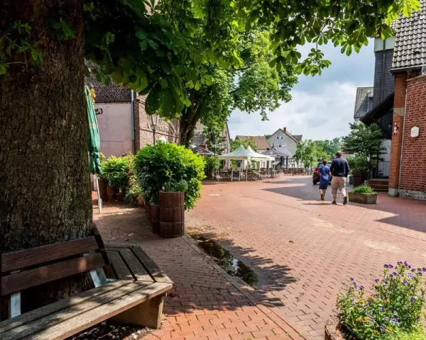 Ein lauschiger Kirchplatz in Recklinghausen mit Kopfsteinpflaster, Bäumen und Sitzgelegenheiten. Ein Paar spaziert vorbei an Cafés mit Außenbestuhlung.