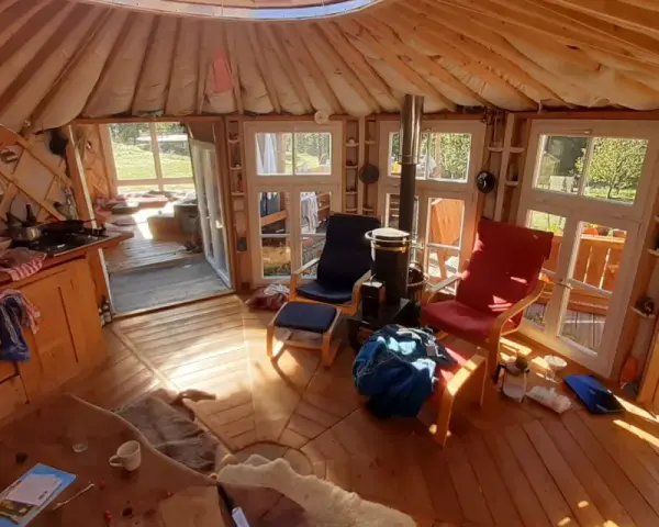 Gemütliches Inneres einer Jurte als Wohnhaus mit Holzofen, Küche und Sitzgelegenheiten. Sonnenlicht fällt durch die Fenster.