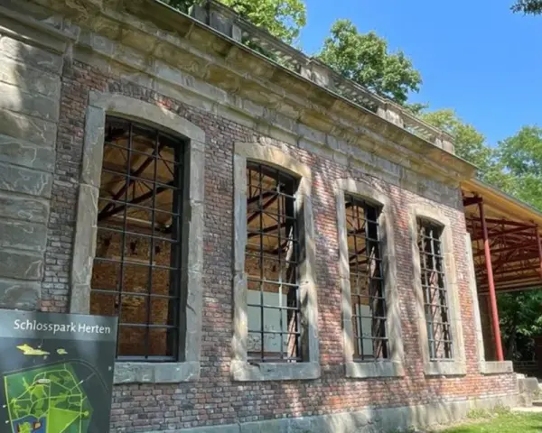 Die Orangerie Herten, ein historisches Gebäude mit großen Fensterfronten, steht im Schatten von Bäumen.