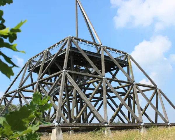 Der Spurwerkturm Waltrop, eine beeindruckende Holzkonstruktion, ragt gegen einen blauen Himmel mit Wolken empor. Grüne Blätter rahmen die Szene ein.
