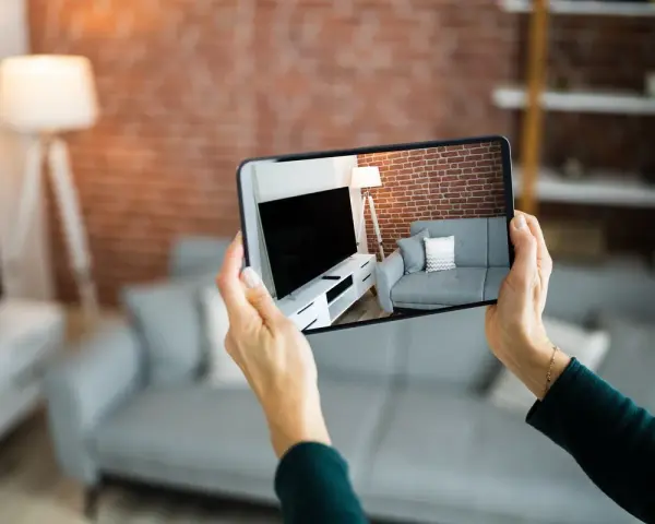 Hände halten Smartphone, das eine Wohnung mit Sofa und Fernseher zeigt. Darf ein Makler Fotos von meiner Wohnung machen? Ja, für die Vermarktung.