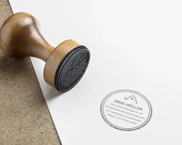 Holzstempel liegt auf Papier neben einem Stempelabdruck von Jens Müller, Sachverständiger für Immobilienbewertung. Infos zu Immobilienpreise Hamm.