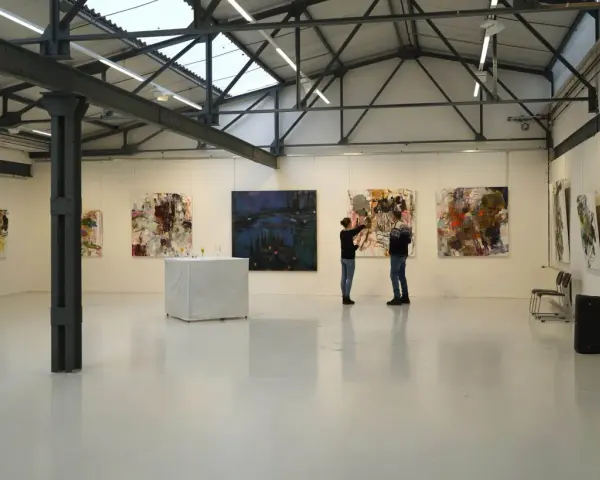 Zwei Personen betrachten Kunstwerke in der Kunsthalle Burkamp. Abstrakte Gemälde hängen an den weißen Wänden, ein Tisch mit Gläsern steht in der Mitte.