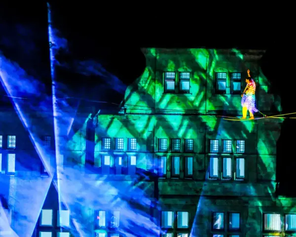 Ein Artist balanciert auf einem Hochseil vor einem Gebäude, das mit grünen Lichtmustern beleuchtet ist. Blaue Lichtstrahlen durchziehen die Szene. Das ist ein Highlight, wann ist Recklinghausen leuchtet!