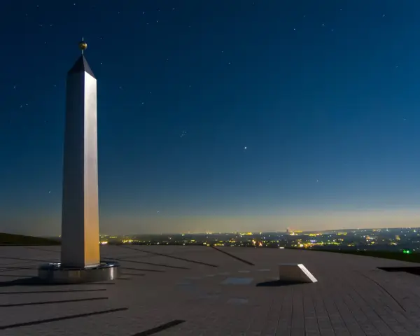 Die Sonnenuhr in Herten ragt unter einem sternenklaren Nachthimmel empor. Ein glänzender Obelisk wirft lange Schatten auf die gepflasterte Fläche.