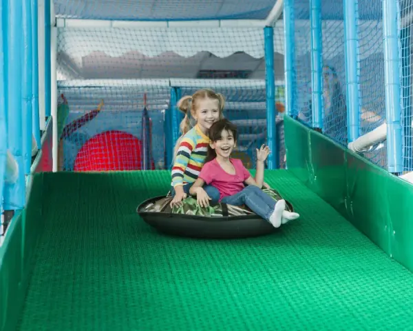 Zwei lachende Mädchen rutschen auf einem Schlauchboot in einem Indoor-Spielplatz.
