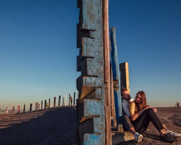 Ein Paar sitzt an der Halde Haniel vor blauen Holzskulpturen. Im Hintergrund eine Reihe bunter Holzpfähle.
