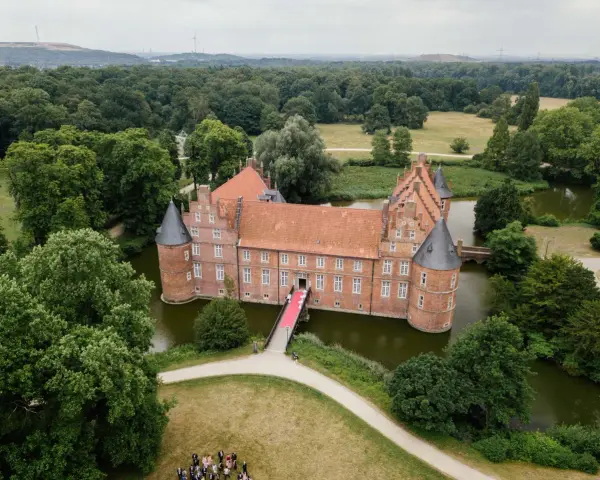 Schloss Recklinghausen, ein imposantes Wasserschloss, umgeben von grünen Bäumen und einem Teich. Eine rote Teppich führt zur Eingangstür.